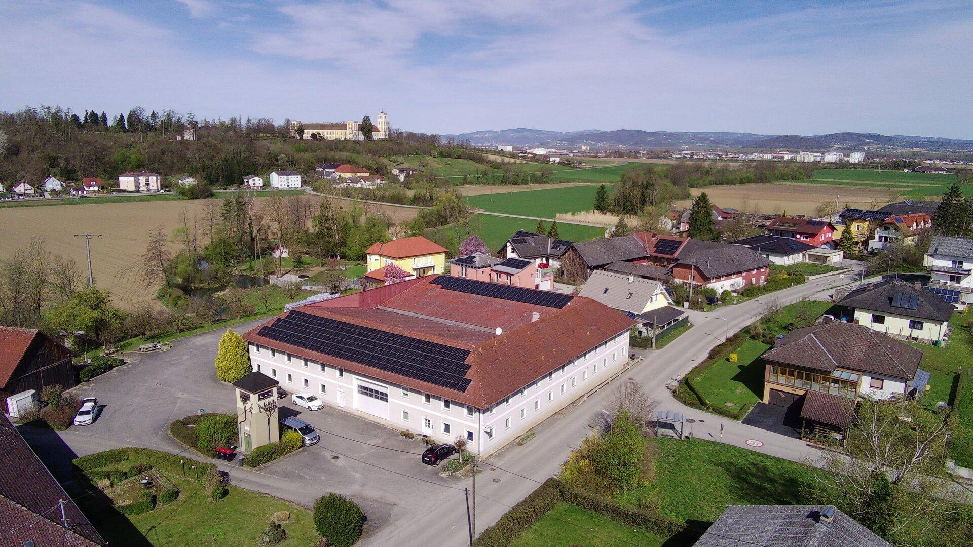 Weitläufige Luftaufnahme des Gewerbegebäudes mit Solaranlagen, umgeben von Wohngebieten und grünen Feldern.