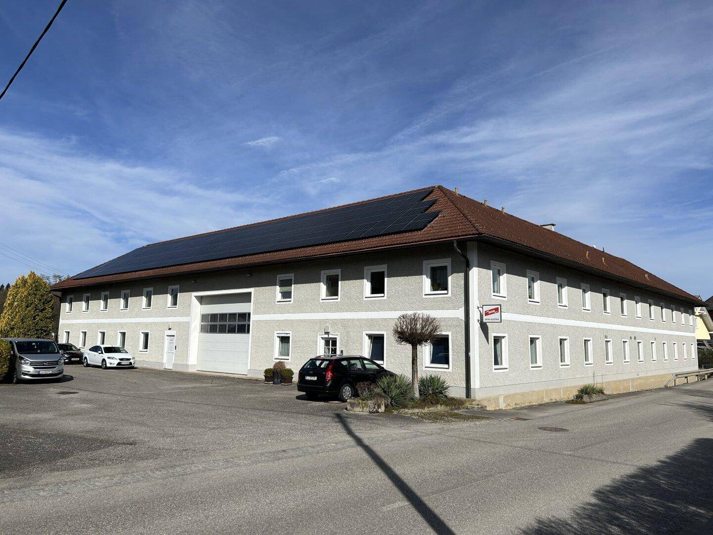 Ansicht des großen Gebäudes mit Solaranlagen auf dem Dach und einem geräumigen Parkplatz davor unter blauem Himmel.