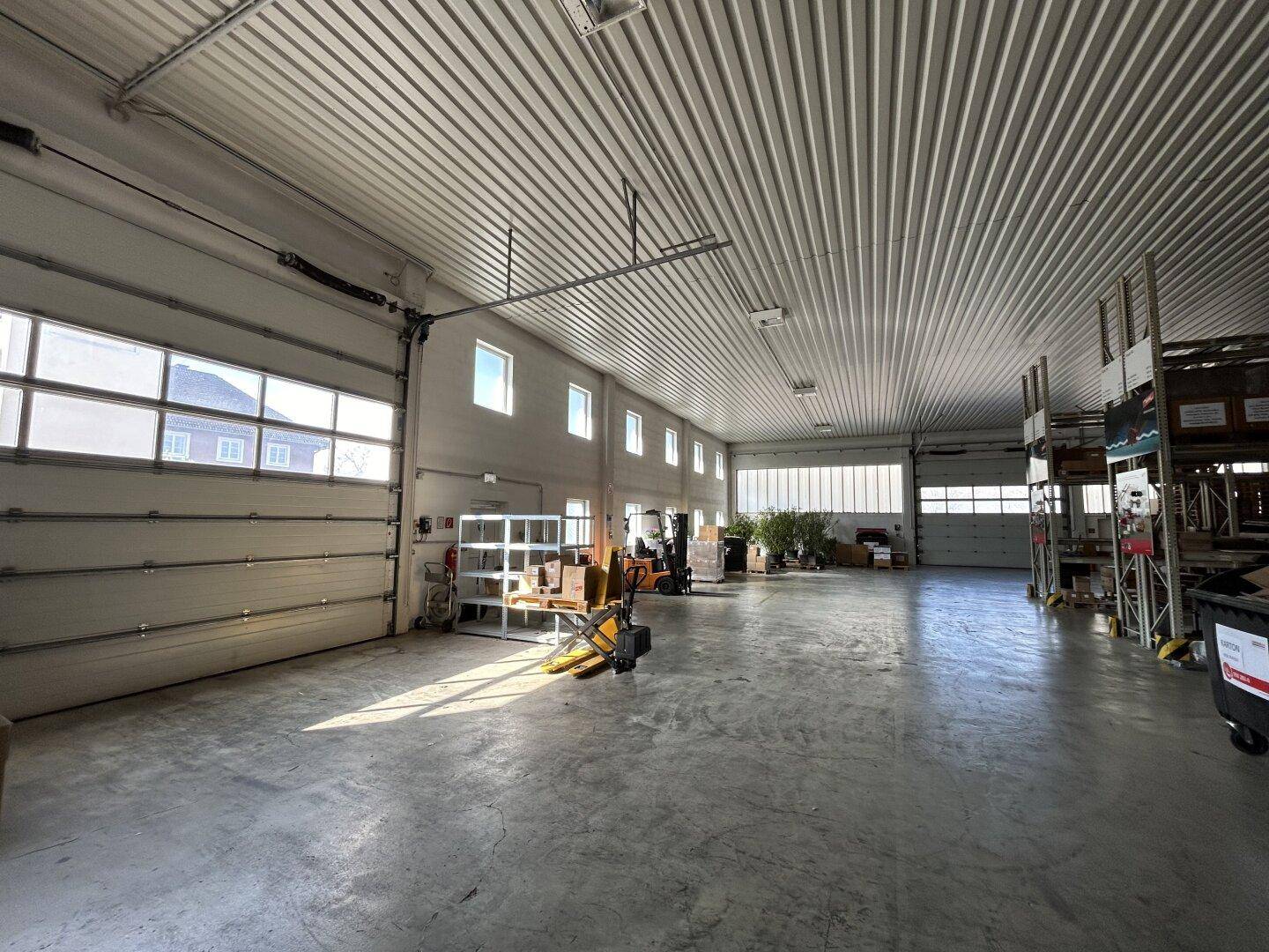 Geräumige Lagerhalle mit hohen Decken, Betonboden und großen Rolltoren, die viel Tageslicht hereinlassen.