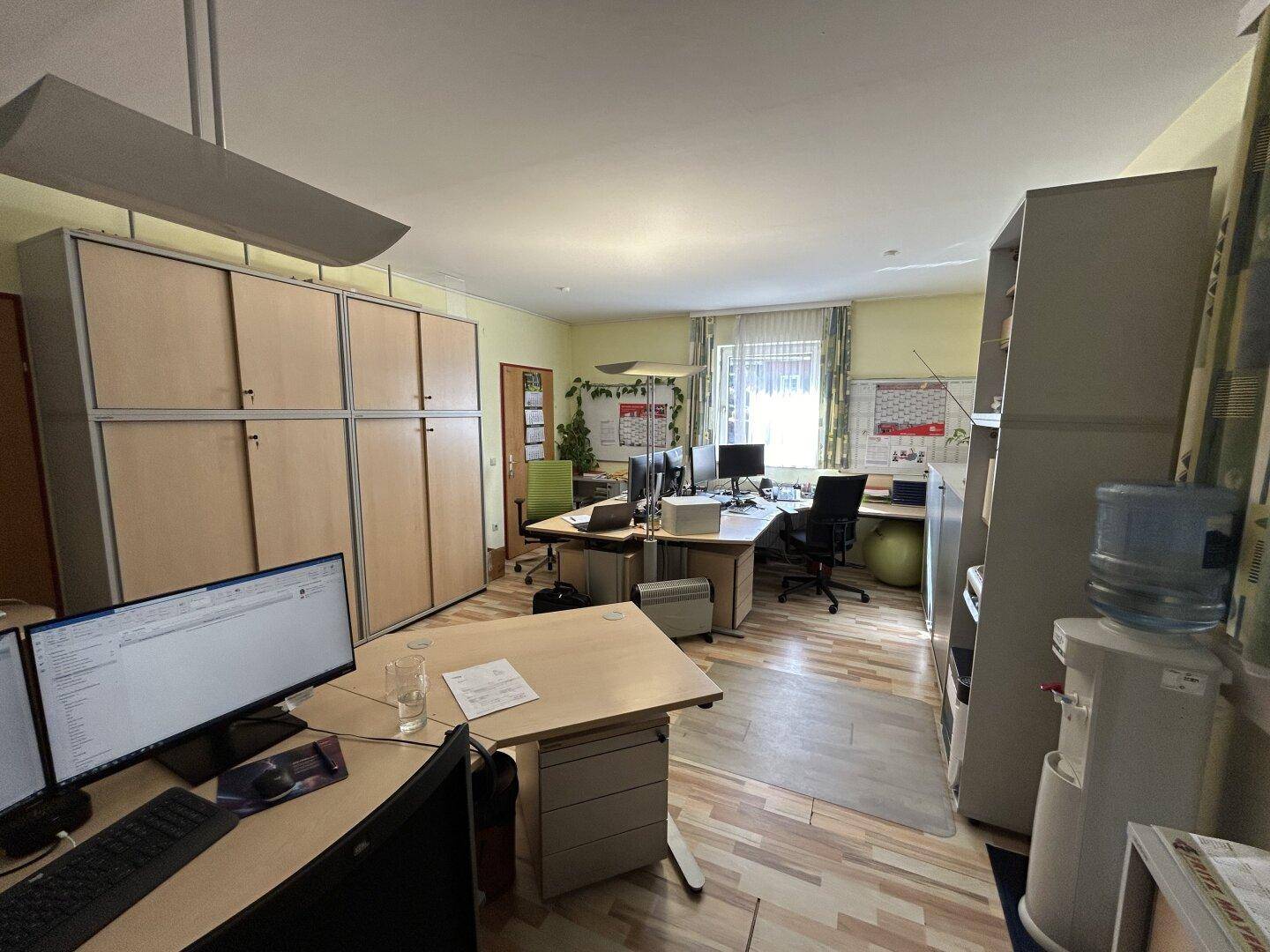 Großes Büro mit mehreren Schreibtischen, Einbauschränken und einem Fenster, das Tageslicht hereinlässt.