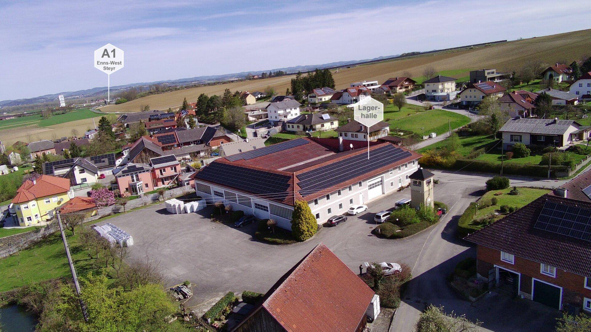 Luftaufnahme eines großen Gebäudes mit Solaranlagen auf dem Dach, umgeben von ländlicher Bebauung und grünen Feldern.
