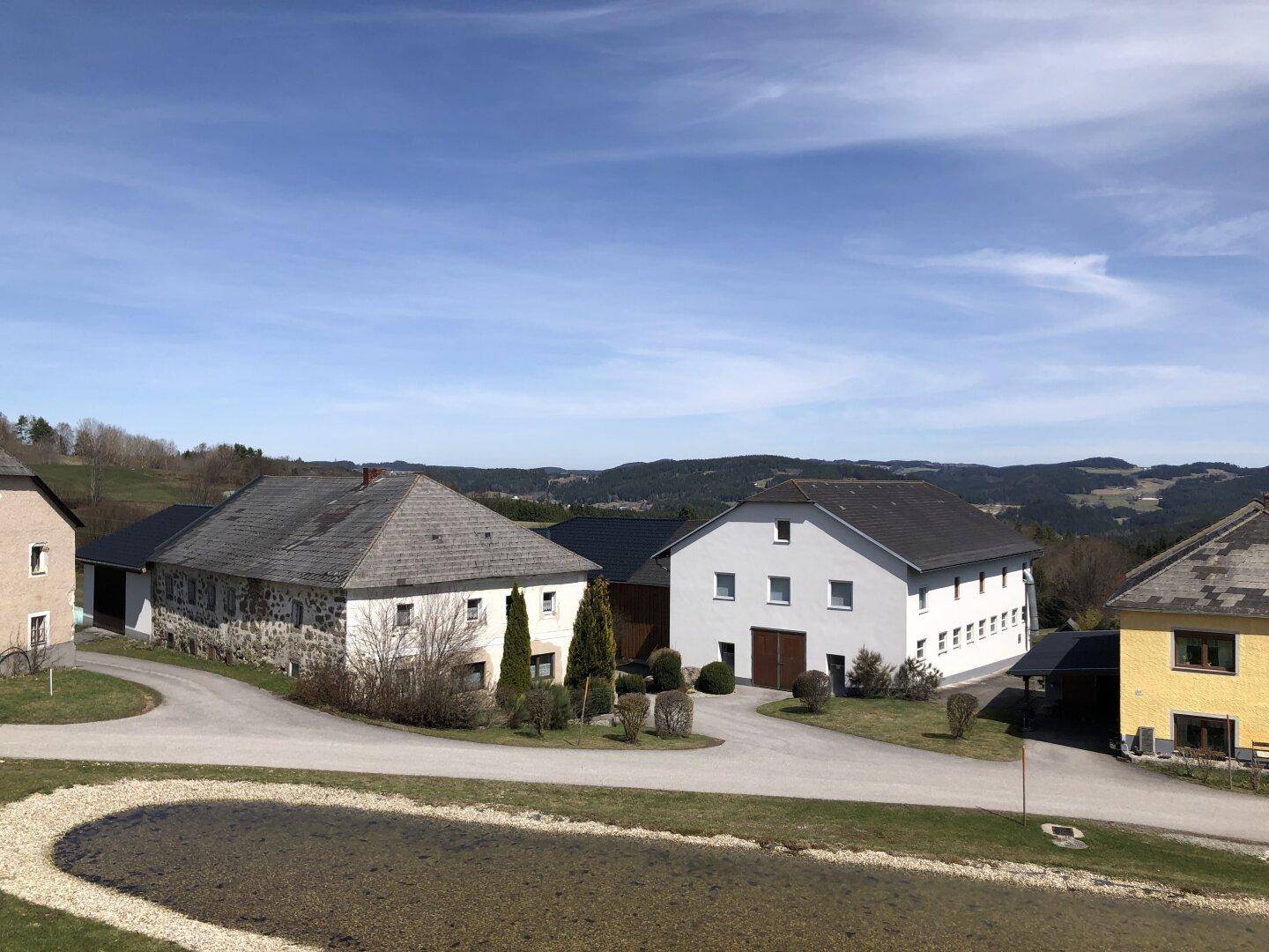 Panoramablick auf ein Bauernhofensemble mit Zufahrt und weitreichender Hügellandschaft im Hintergrund.