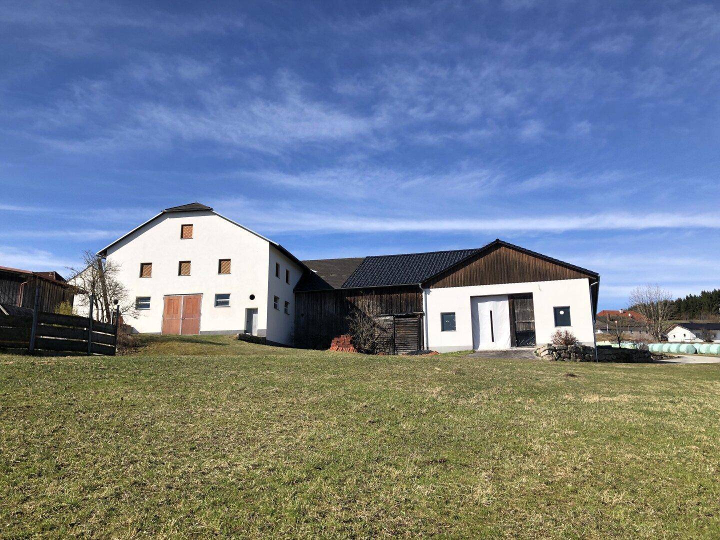 Großes weißes Bauernhaus mit dunklem Dach und angrenzendem Holzbau, umgeben von weitläufigen Grünflächen.