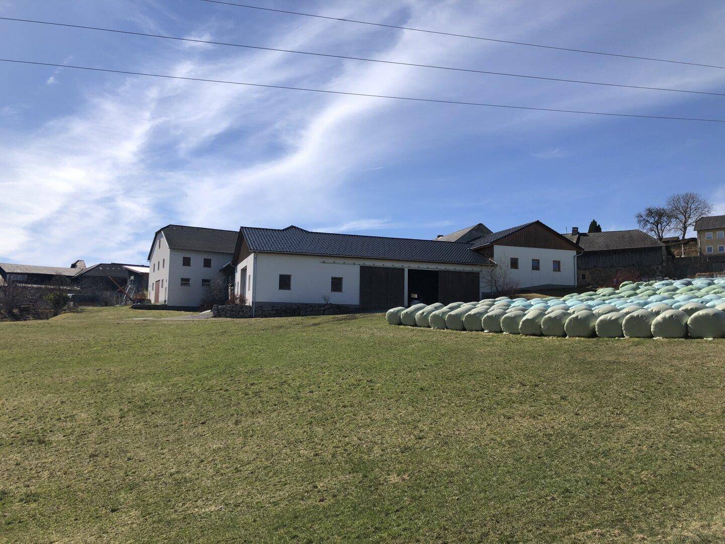 Weitläufiges Bauernhofgelände mit mehreren Gebäuden, grüner Wiese und gestapelten Heuballen unter blauem Himmel.