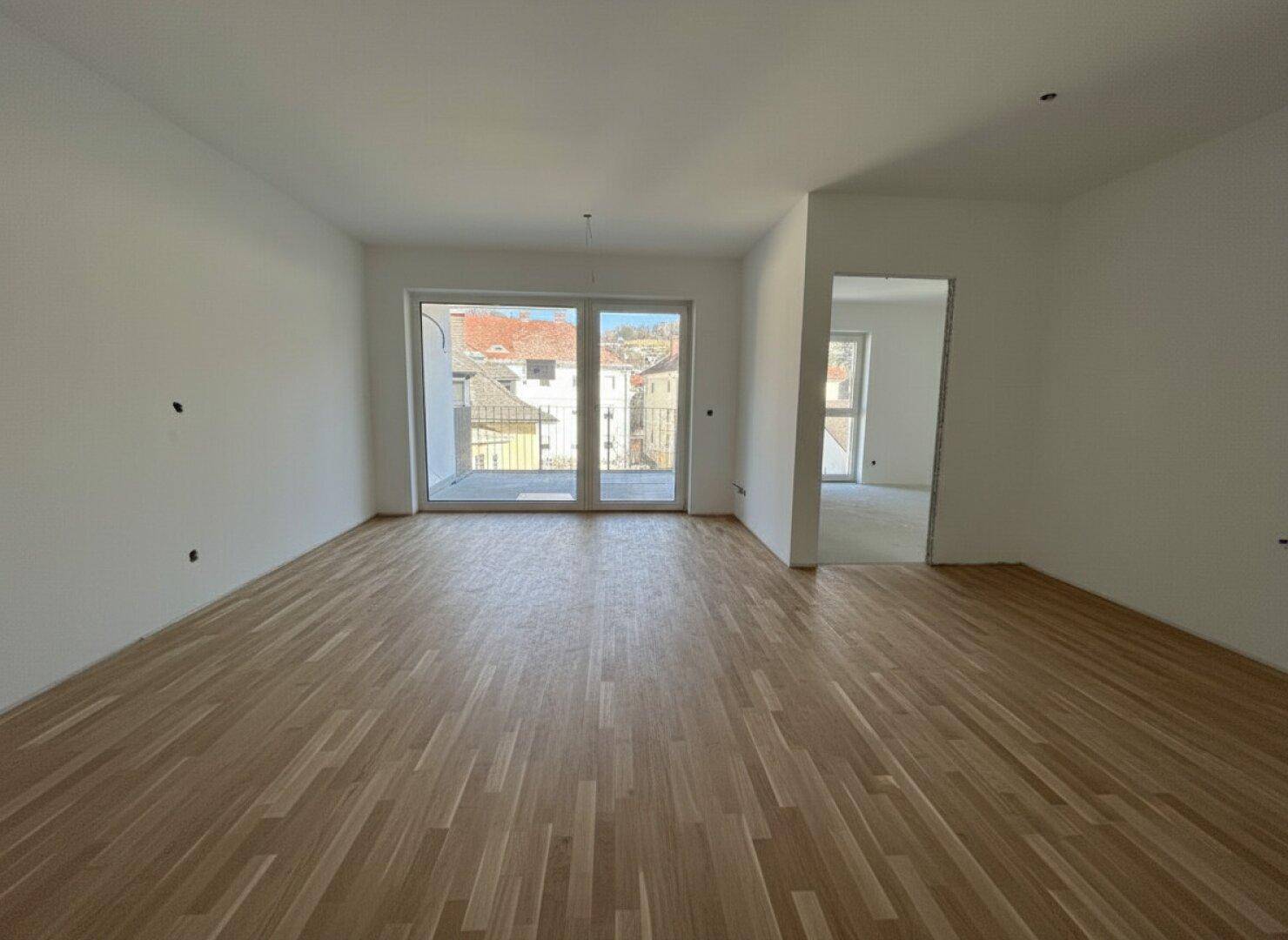 Heller, geräumiger Wohnbereich mit neuem Parkettboden, weißen Wänden und Zugang zu einem Balkon mit Stadtblick.