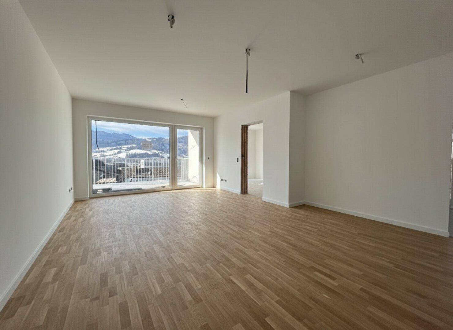 Heller, moderner Wohnraum mit hochwertigem Parkettboden, großen Fenstern und direktem Zugang zu einem Balkon mit Bergblick.