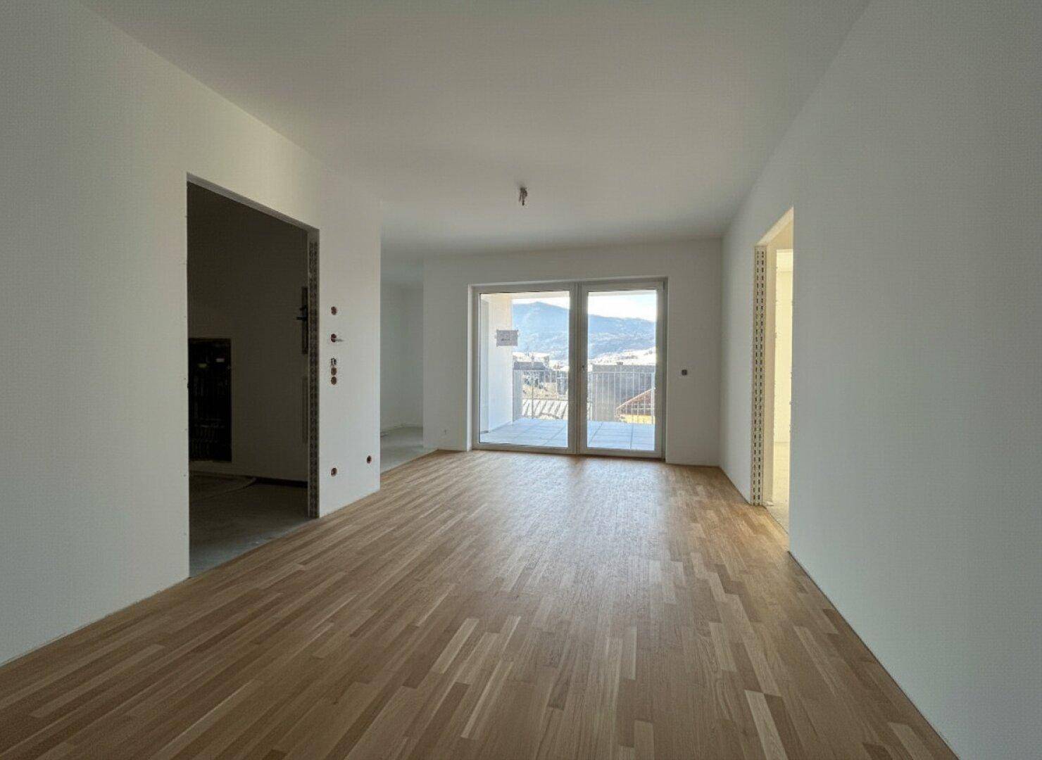 Heller, leerer Raum mit neuem Parkettboden, großen Fenstern und Zugang zu einer Terrasse mit Bergblick.