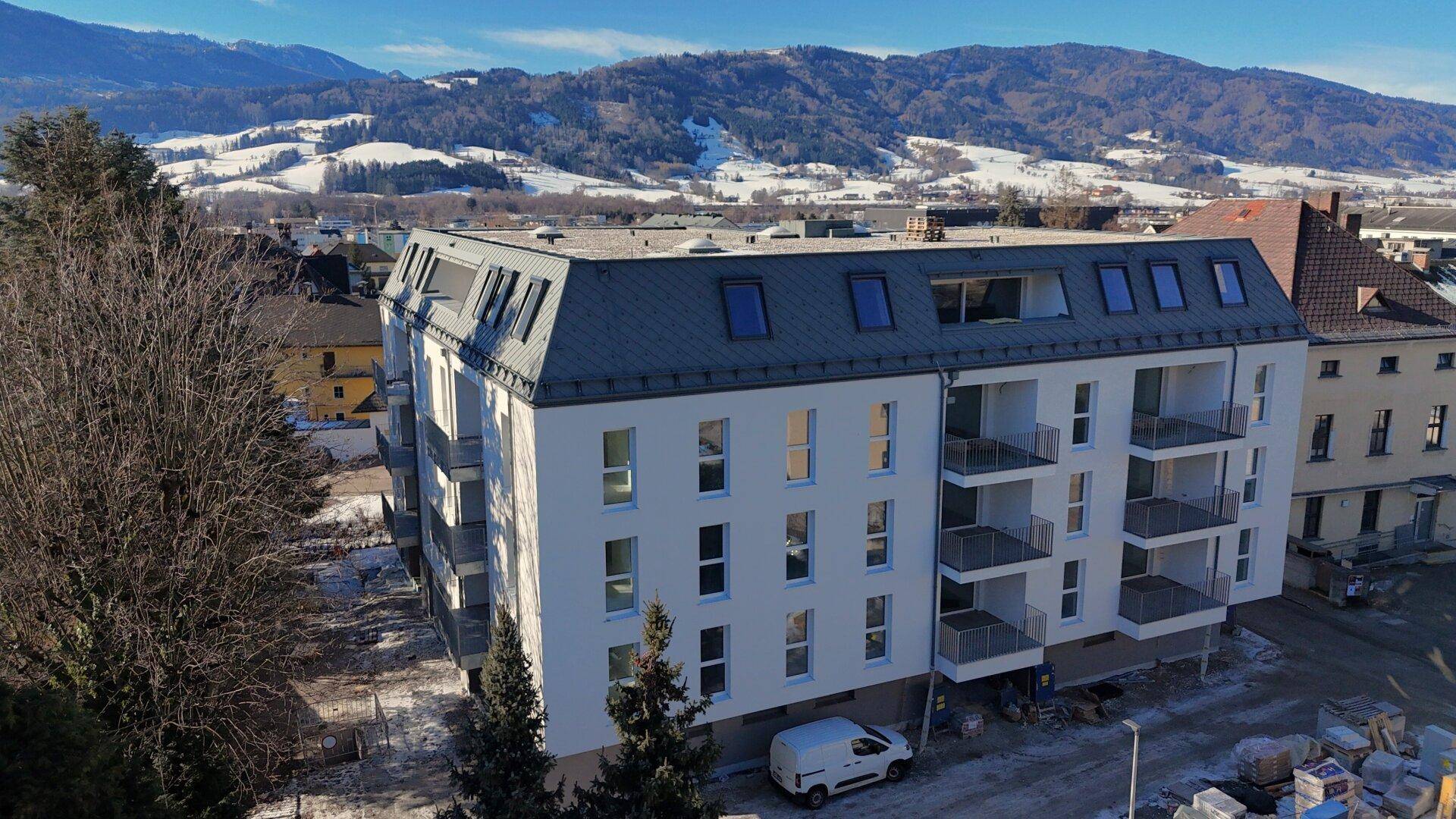 Modernes Wohngebäude mit Balkonen und Mansarddach, umgeben von schneebedeckten Hügeln.