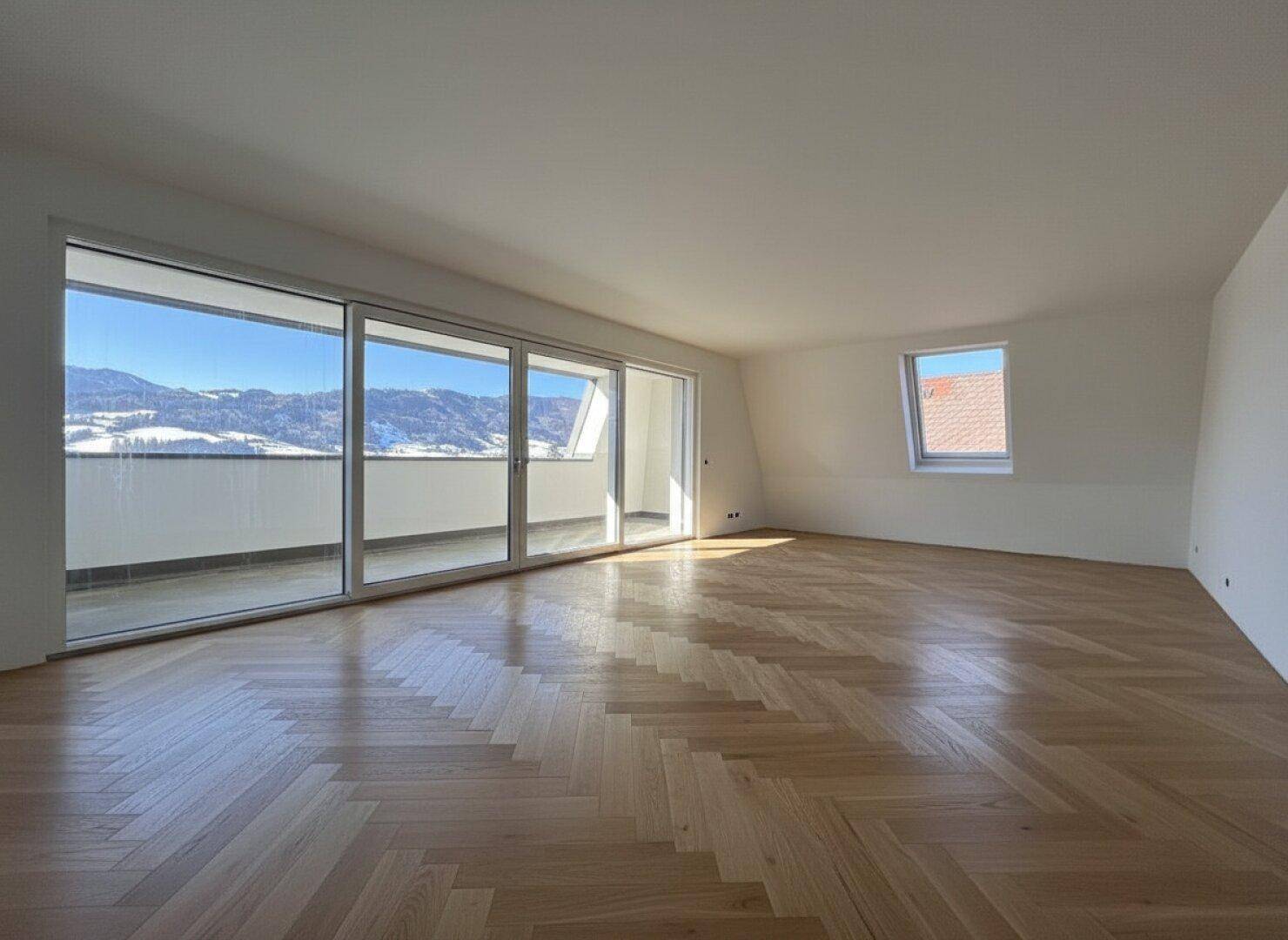 Helles, leeres Zimmer mit Fischgrätparkett und großen Fenstern mit Bergblick.