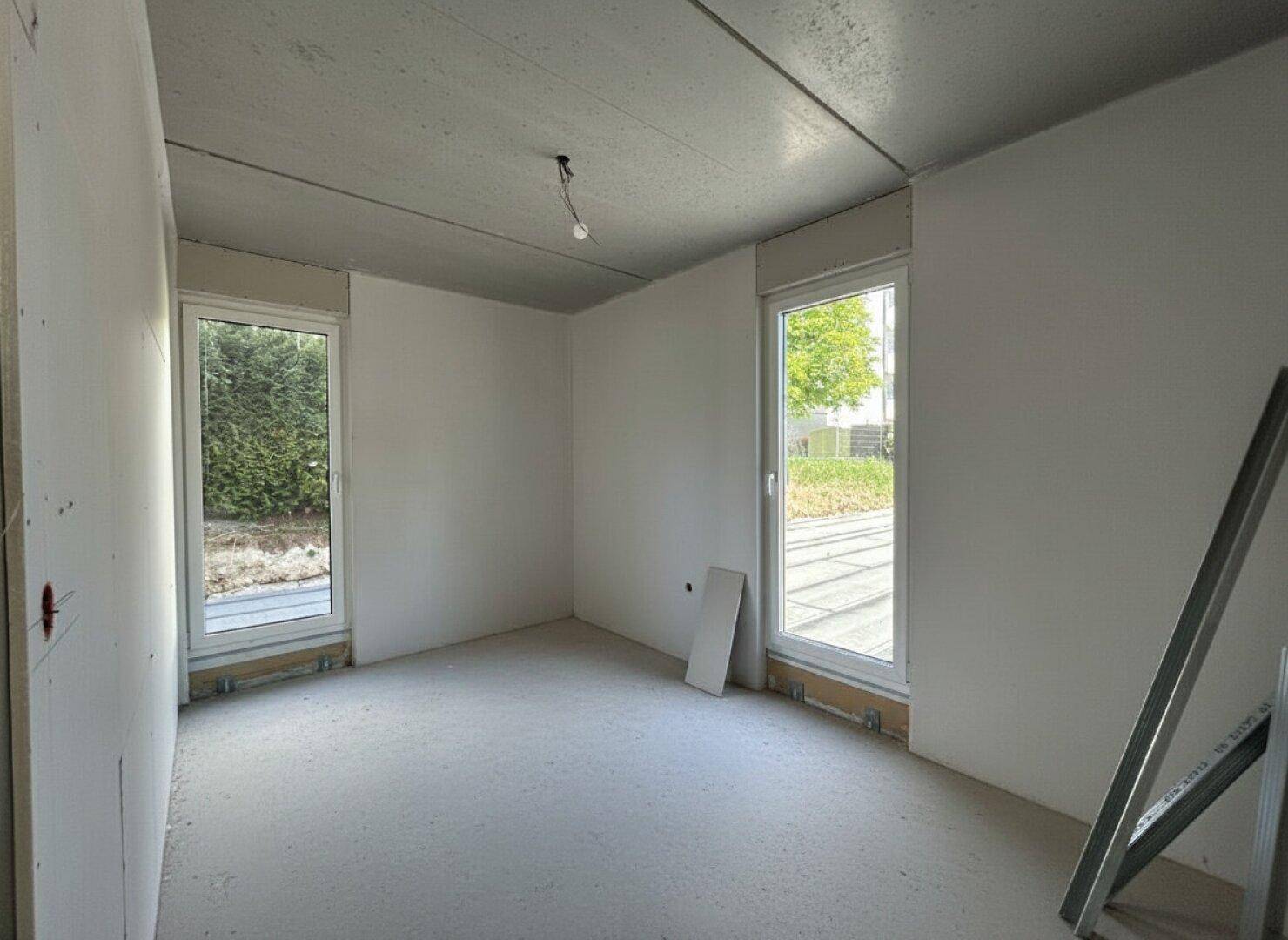 Helles Zimmer im Rohbau mit zwei Fenstern und Blick auf die Umgebung.
