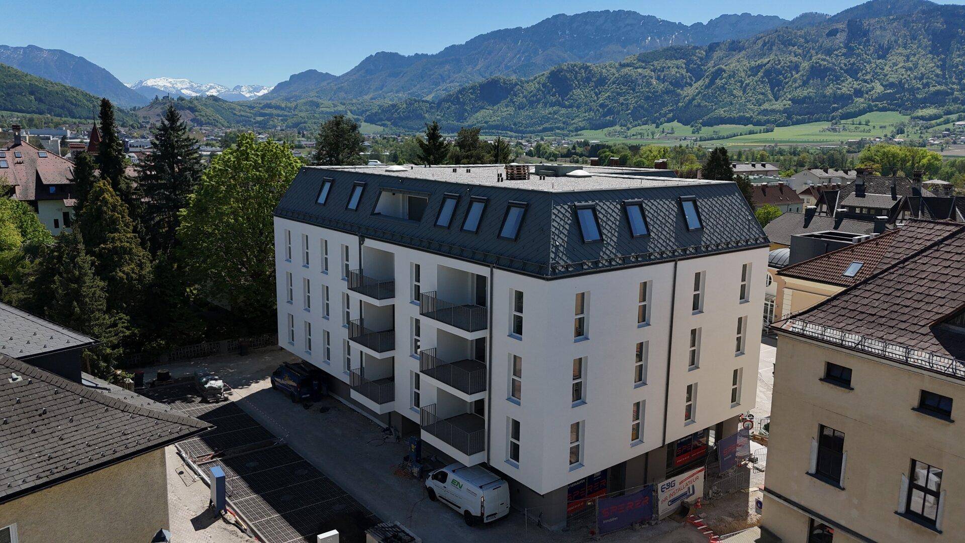 Neubau mit weißer Fassade und Balkonen, umgeben von älteren Gebäuden und einer Bergkulisse.
