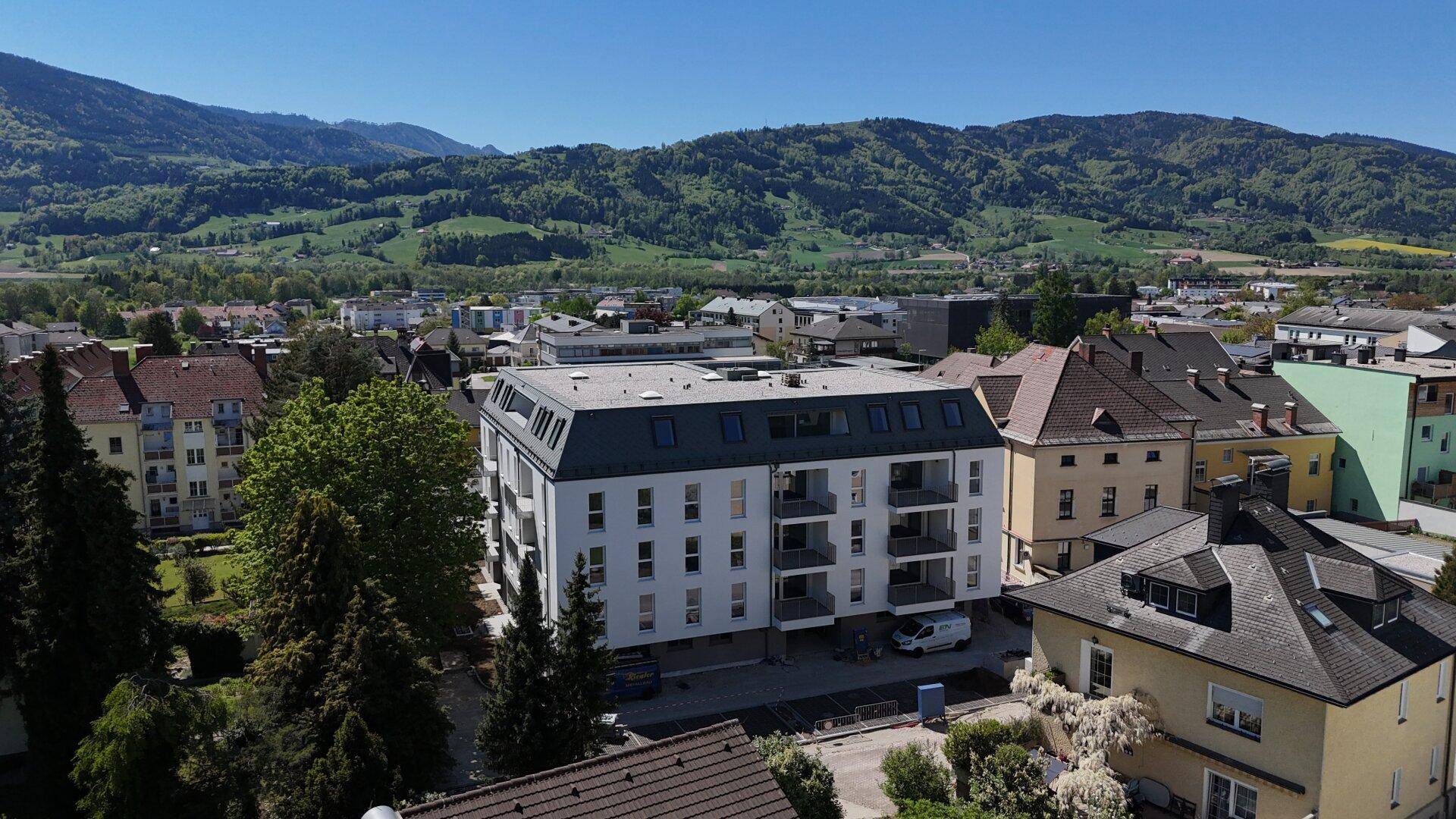 Modernes Wohngebäude mit Mansarddach und Balkonen, eingebettet in eine grüne Landschaft mit Bergen.