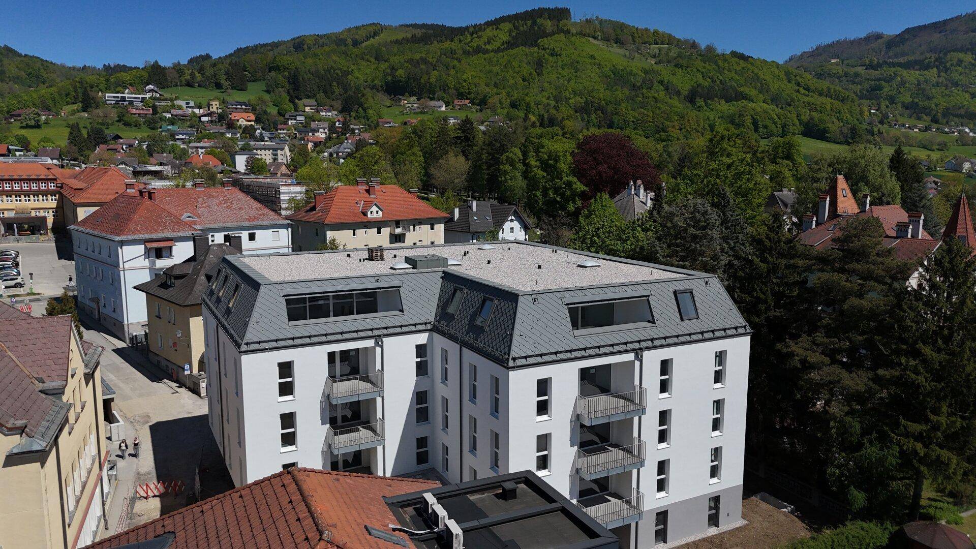 Modernes Wohngebäude mit Balkonen und Mansarddach, umgeben von grünen Hügeln und älteren Häusern.