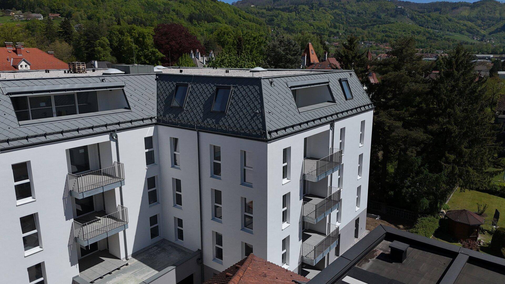 Moderne Wohnanlage mit Balkonen und Mansarddach, umgeben von grünen Hügeln.