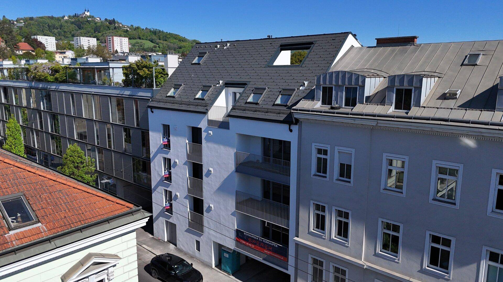 Ansicht der modernen Gebäudefassade mit Balkonen und Dachfenstern, umgeben von älteren Gebäuden und grünen Hügeln.