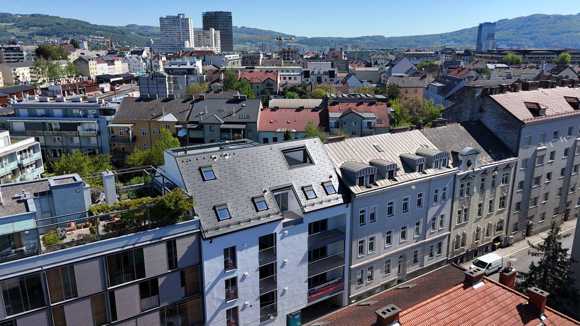 Luftaufnahme der Stadt mit Wohngebäuden und grünen Hügeln im Hintergrund unter blauem Himmel.