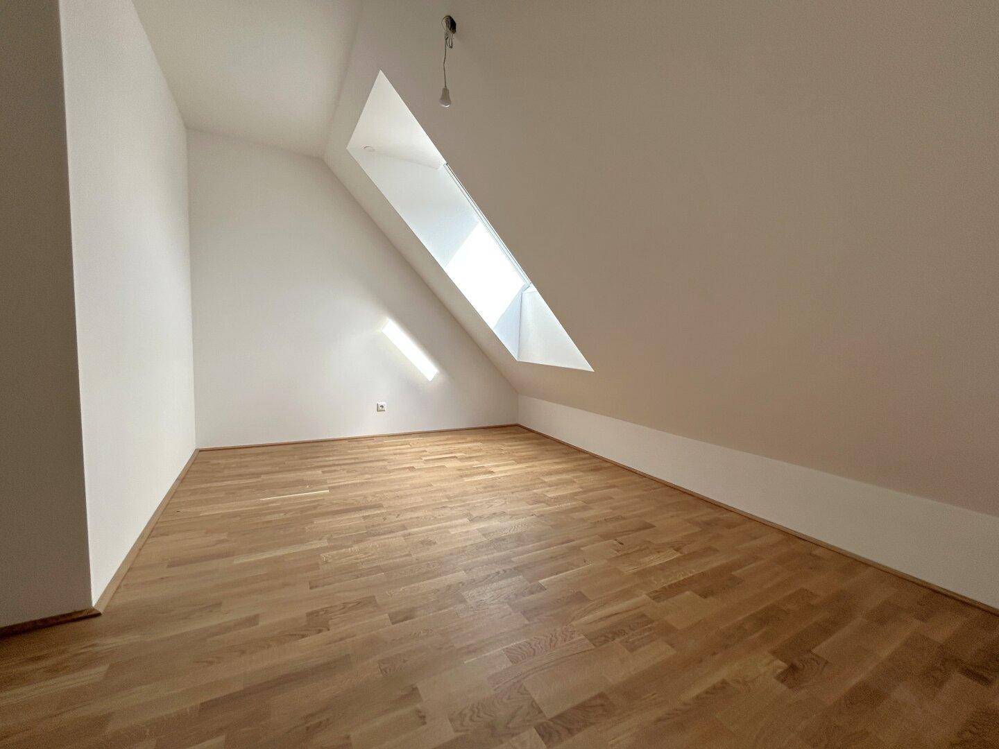 Leerer Raum mit Holzboden und schrägem Dachfenster, das viel Tageslicht hereinlässt.