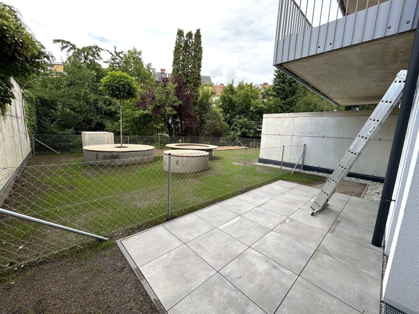 Außenbereich mit gepflasterter Terrasse, Rasenfläche und erhöhten Pflanzbeeten, umgeben von Grün.