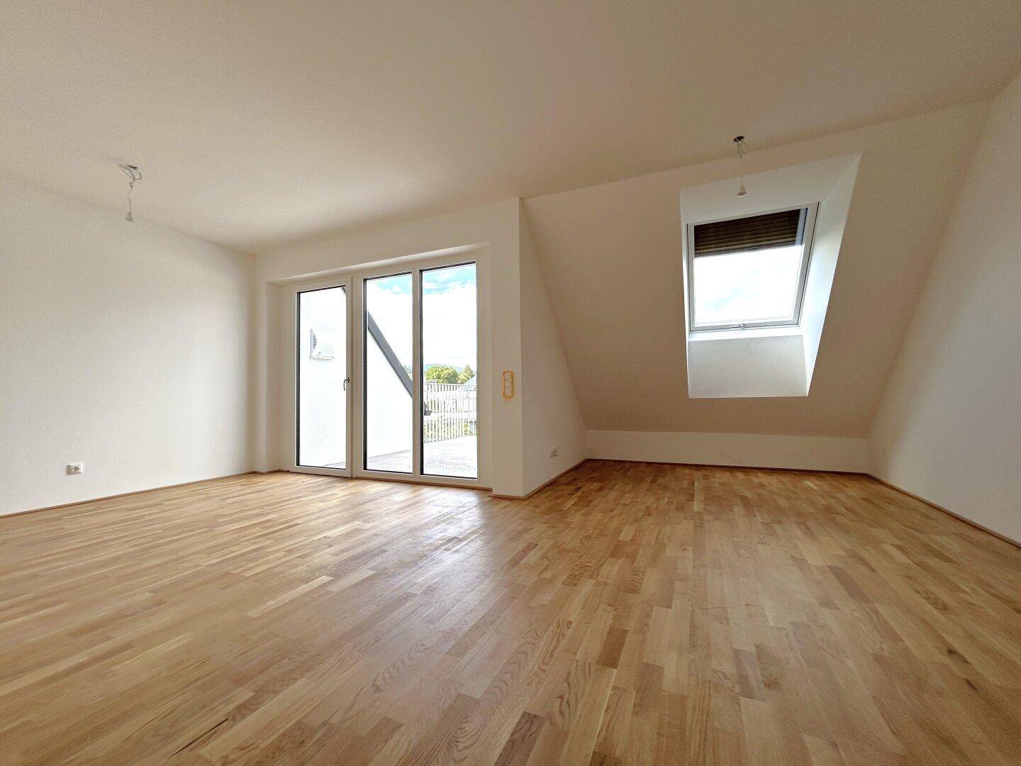 Geräumiges Zimmer mit Holzboden, großen Fenstern und einem Dachfenster mit Rollladen.