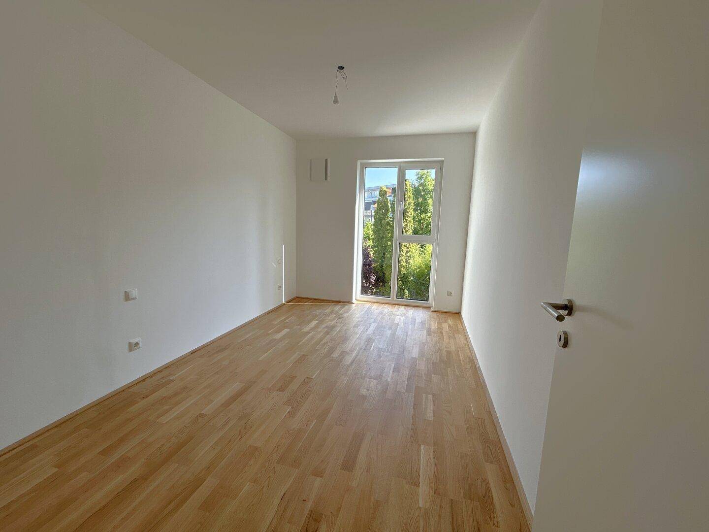 Lichtdurchflutetes Zimmer mit Parkettboden und Fenster mit Blick auf Bäume und Natur.