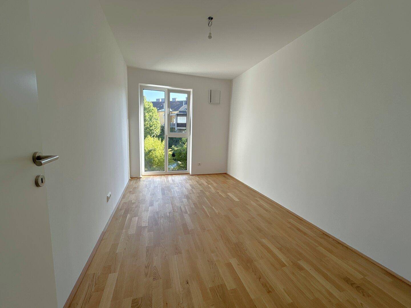 Helles Zimmer mit Holzboden und großem Fenster, das viel Tageslicht hereinlässt und Grünblick bietet.