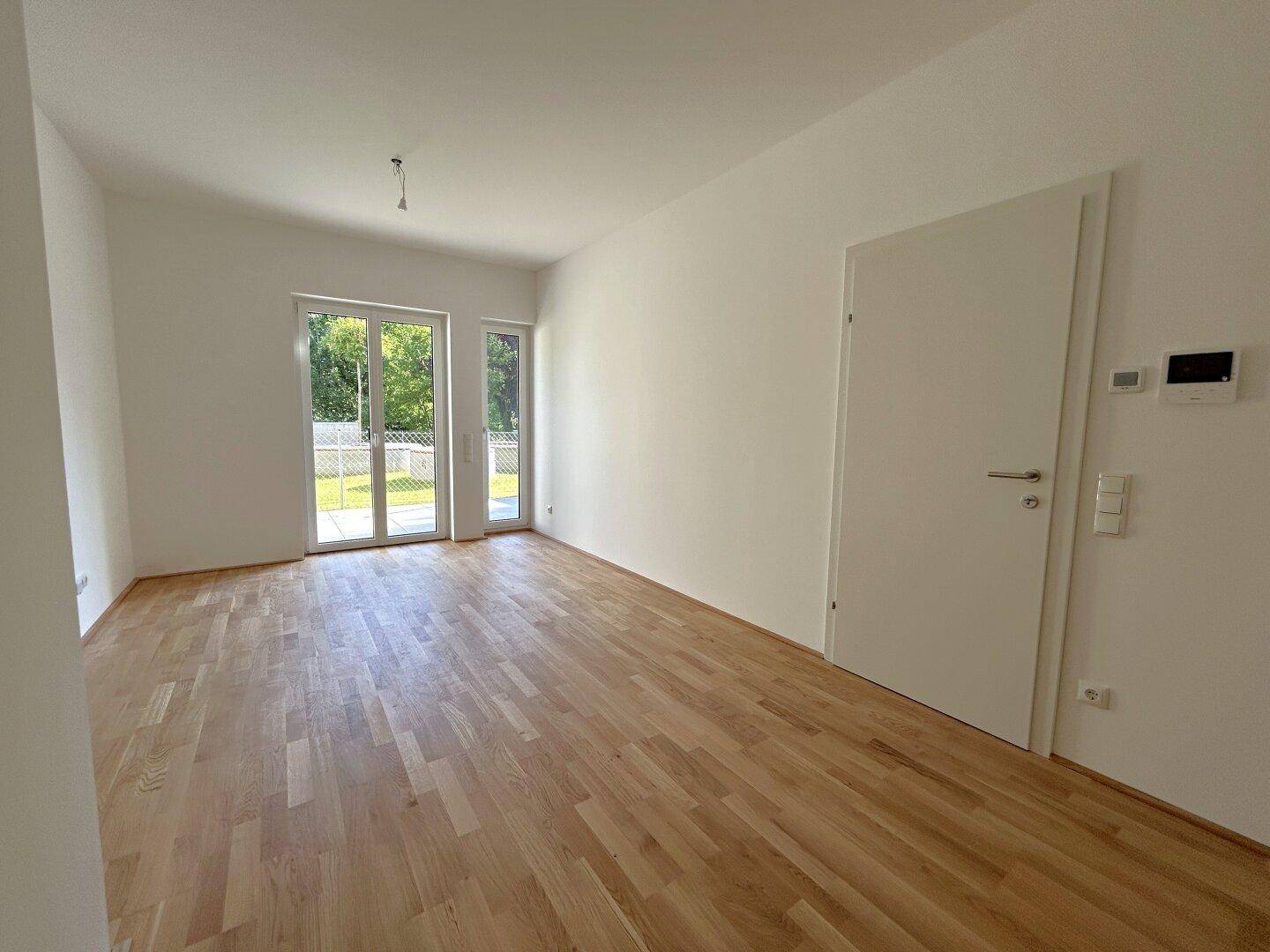 Geräumiges, helles Zimmer mit Holzboden und Zugang zu einem Außenbereich durch Fenstertüren.