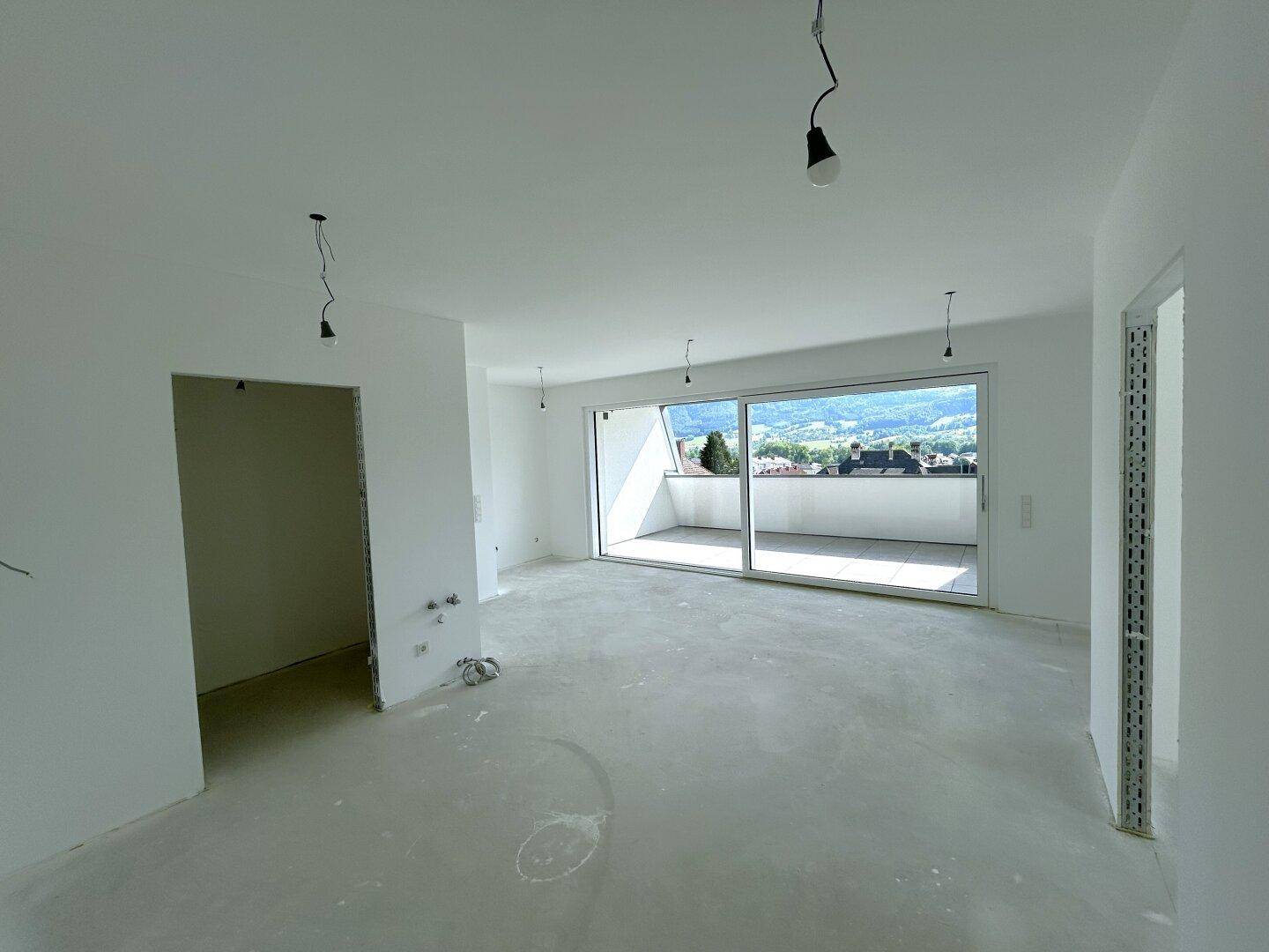 Unfertiger Wohnbereich mit Rohbetonboden und Zugang zu einem Balkon mit Bergblick.