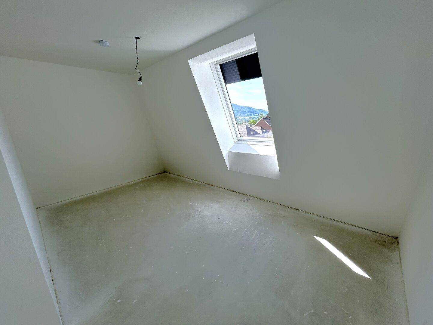 Geräumiges Dachgeschosszimmer mit schrägen Wänden und einem Fenster mit Blick ins Grüne.