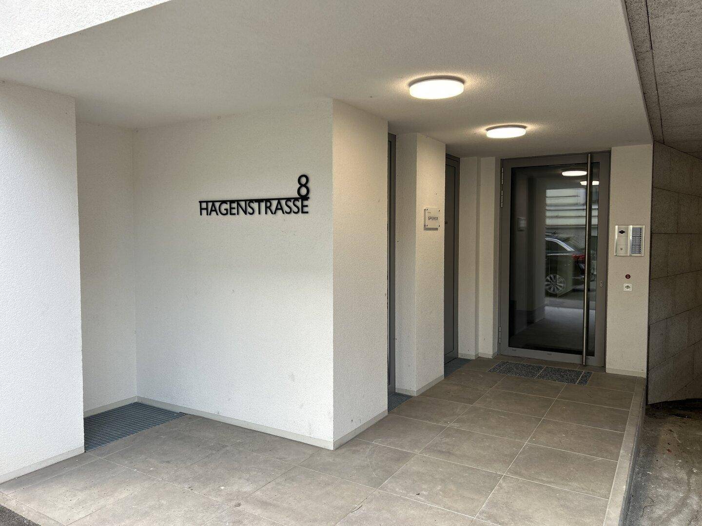 Moderner Eingangsbereich eines Gebäudes mit Fliesenboden, Hausnummer und Gegensprechanlage.