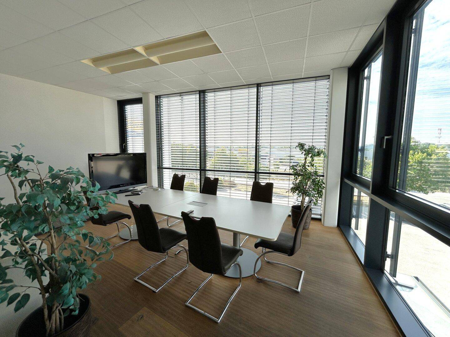 Konferenzraum mit großem Tisch, Stühlen, Fernseher und großen Fenstern mit Jalousien.