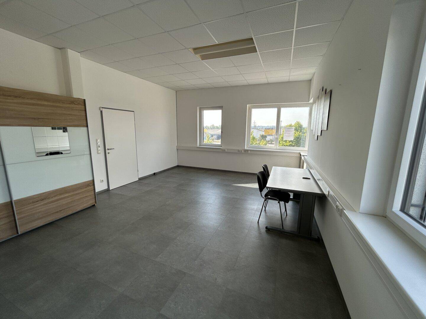 Geräumiges Büro mit grauen Fliesen, einem Schrank und Blick nach draußen durch große Fenster.