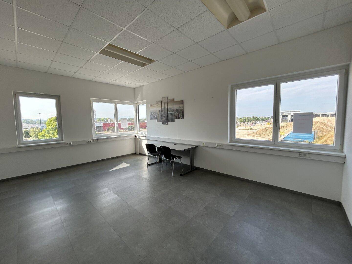 Helles, leeres Büro mit grauen Fliesen, großen Fenstern und einer einfachen Schreibtischgarnitur.
