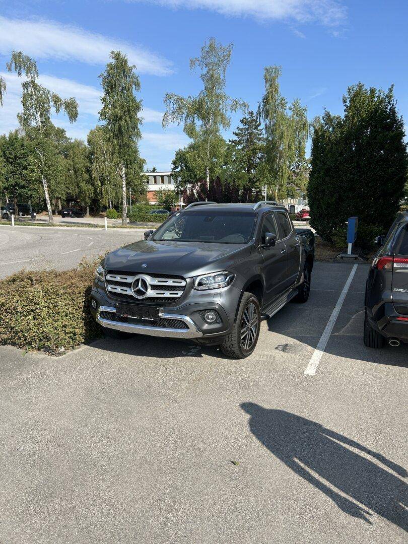 Ein grauer Mercedes-Pickup steht auf einem asphaltierten Parkplatz unter einem sonnigen Himmel.