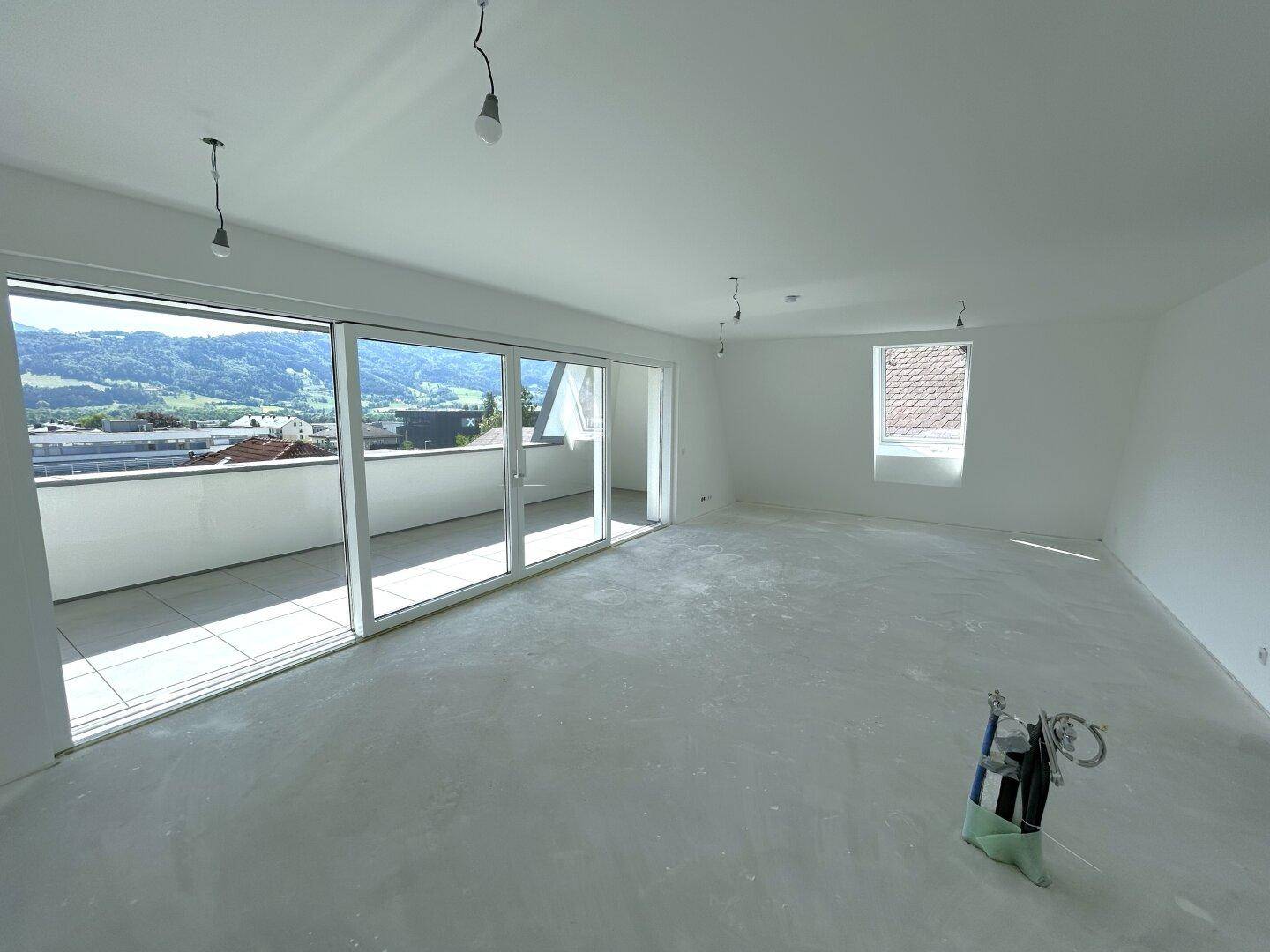 Unfertiger Wohnbereich mit großen Schiebetüren zum Balkon und weitem Blick auf die grüne Berglandschaft.