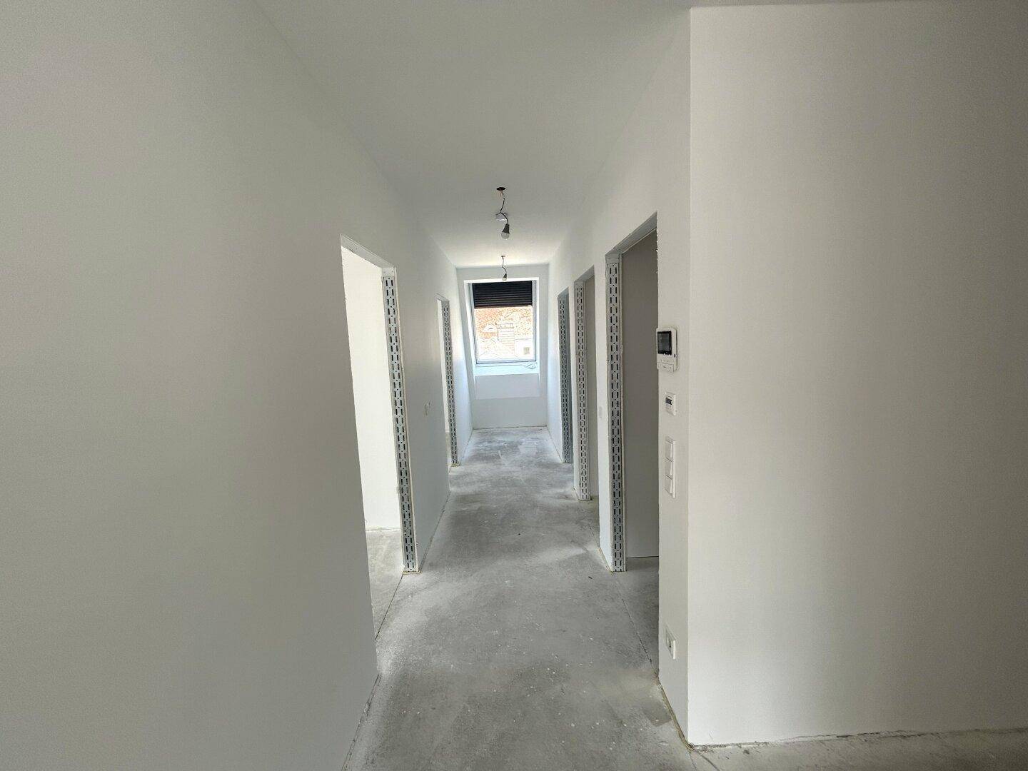 Heller, unfertiger Flur mit Betonboden, weißen Wänden und einem Fenster am Ende für natürliches Licht.