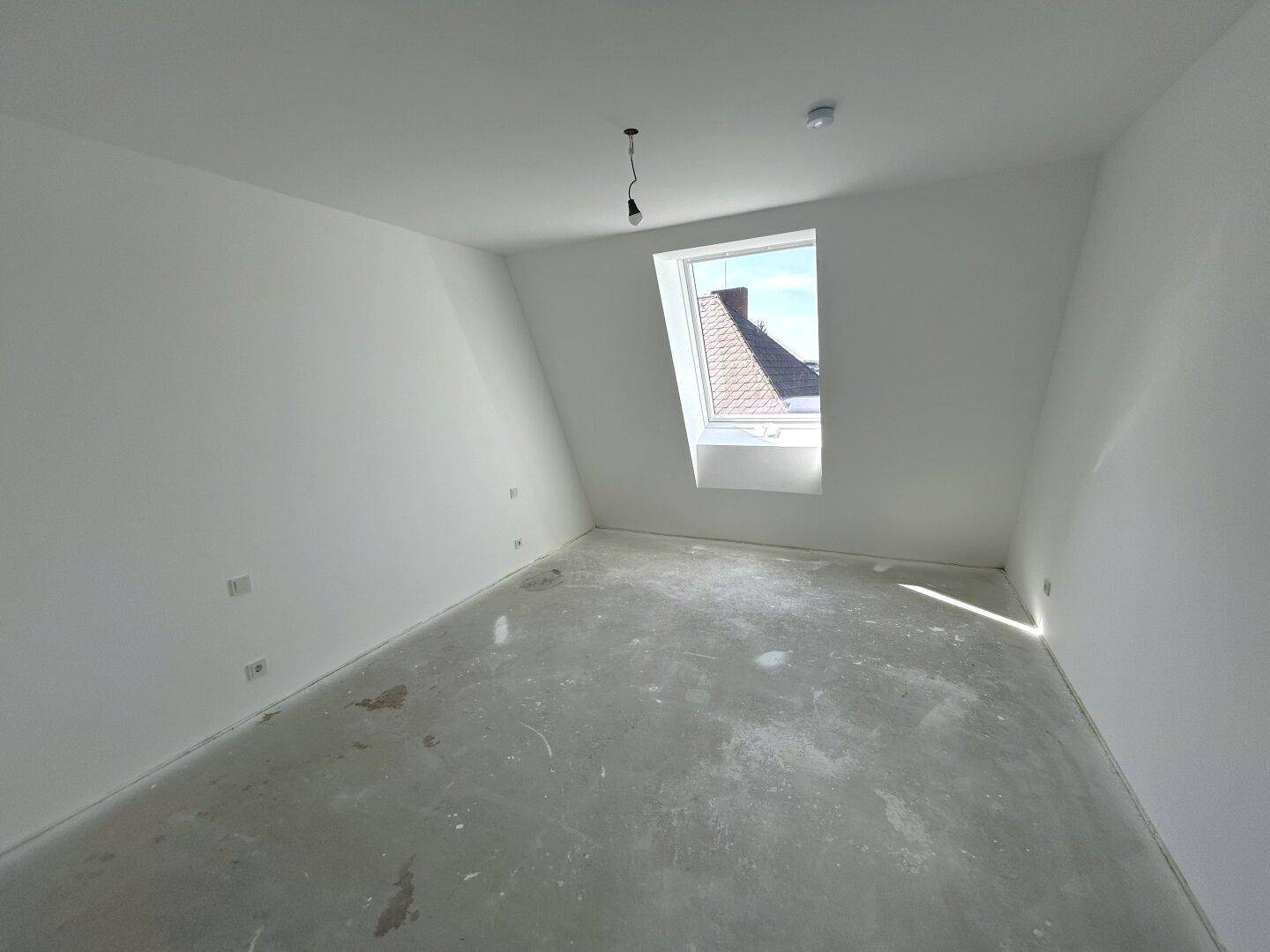 Unfertiges Schlafzimmer im Dachgeschoss mit Betonboden, weißen Wänden und einem Dachfenster.