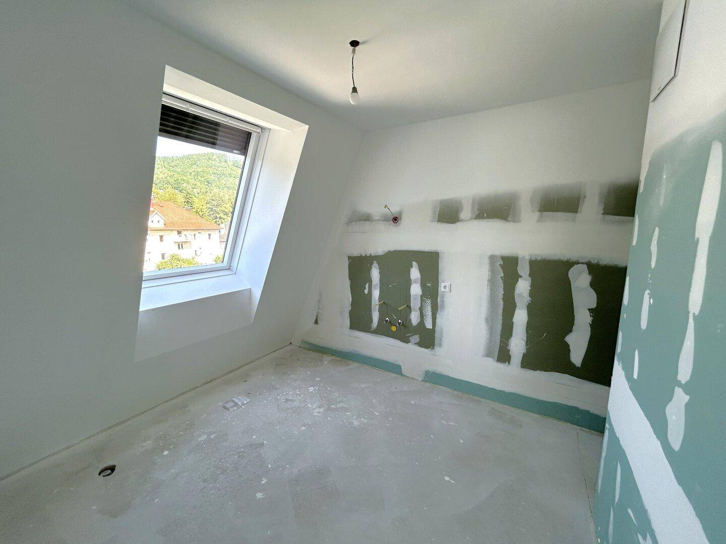 Unfertiger Raum im Dachgeschoss mit Betonboden, Dachfenster und vorbereiteten Installationen für Sanitäranlagen.