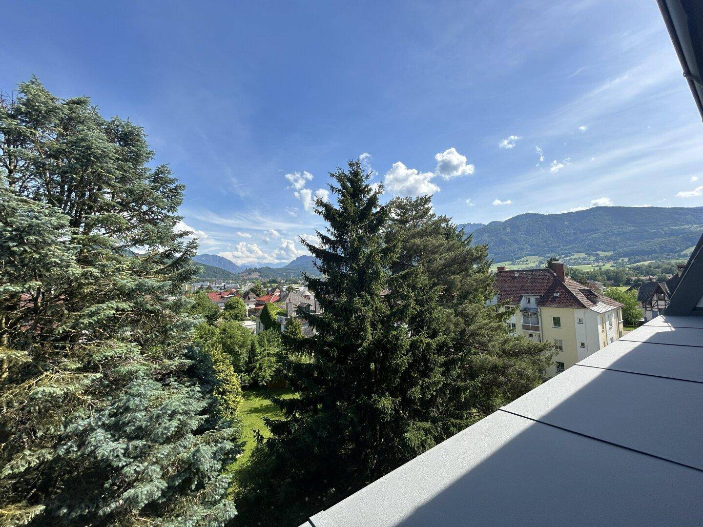 Malerischer Ausblick von einem Balkon auf grüne Bäume, Häuser und die beeindruckende Berglandschaft.