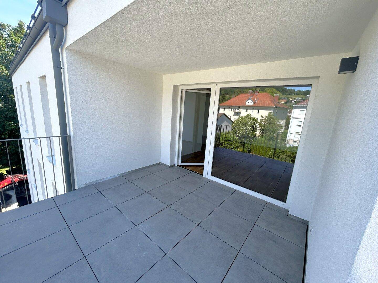 Heller Balkon mit grauen Fliesen und Zugang zum Innenraum, umgeben von einer Wohngegend.