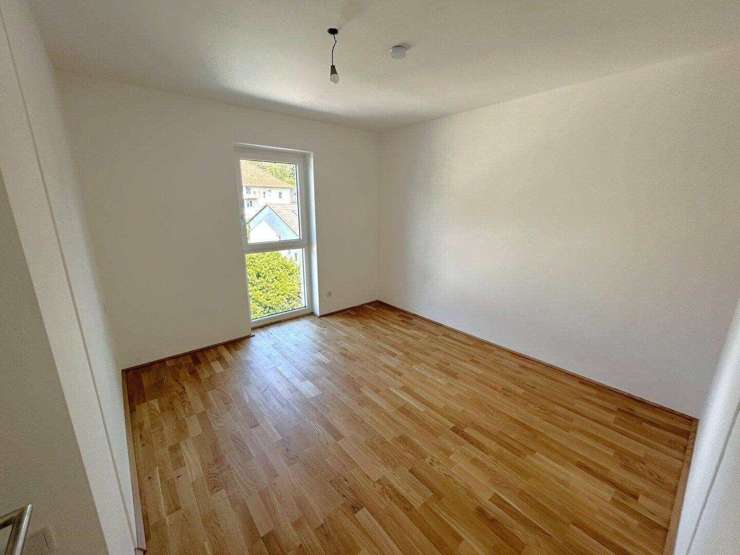 Leeres Schlafzimmer mit Holzboden und einem Fenster, das einen Blick ins Grüne bietet.