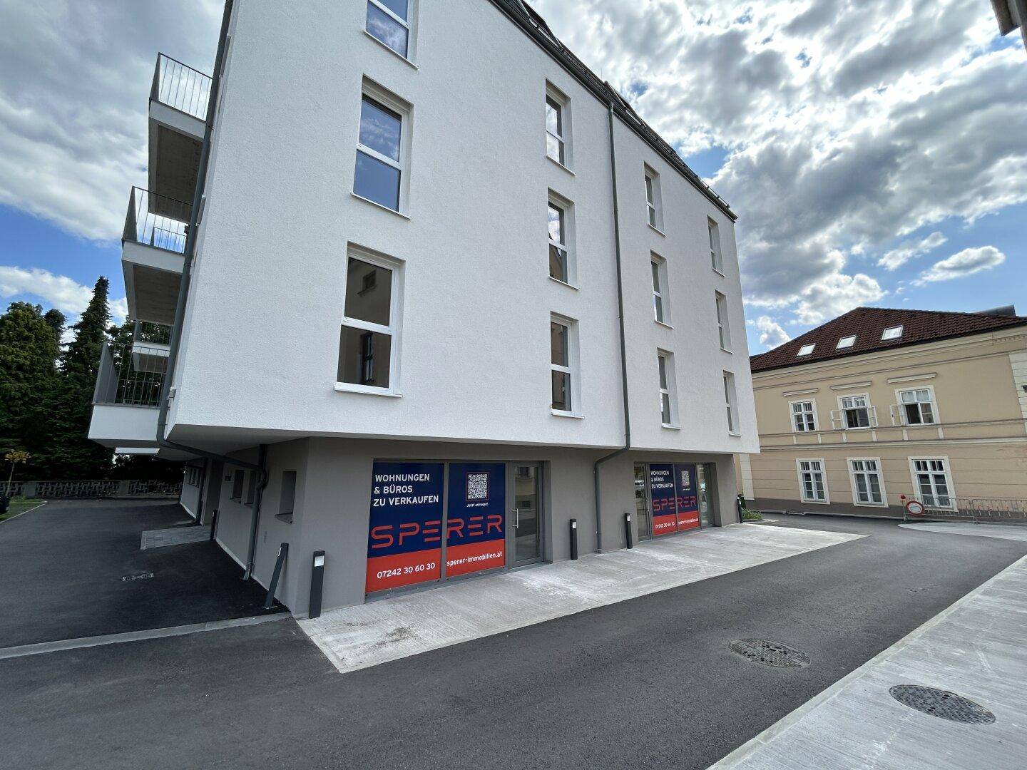 Neubau mit weißer Fassade, Balkonen und Geschäftsflächen im Erdgeschoss unter blauem Himmel.