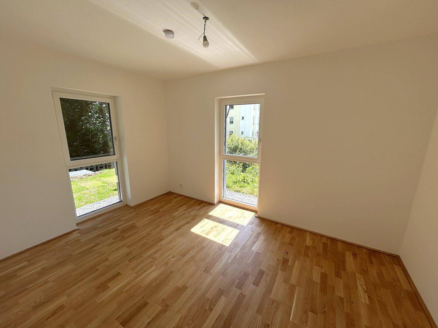 Helles Schlafzimmer mit Holzboden und zwei großen Fenstern mit Blick ins Grüne.