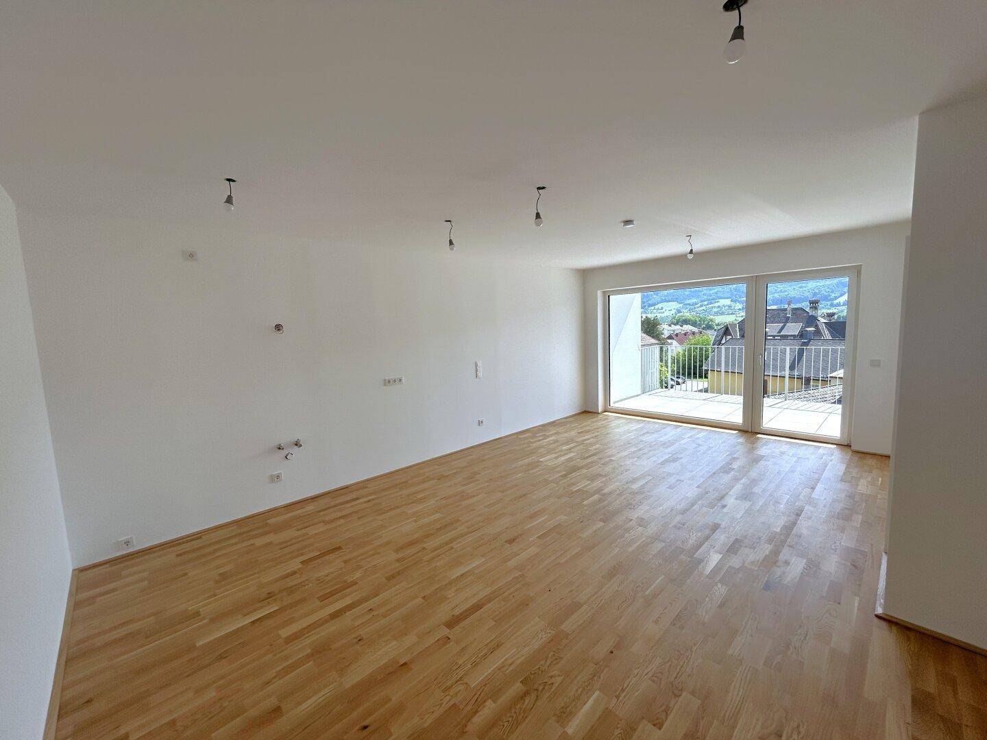 Geräumiges, unmöbliertes Wohnzimmer mit hellem Parkettboden und weitem Blick auf die umliegende grüne Landschaft.