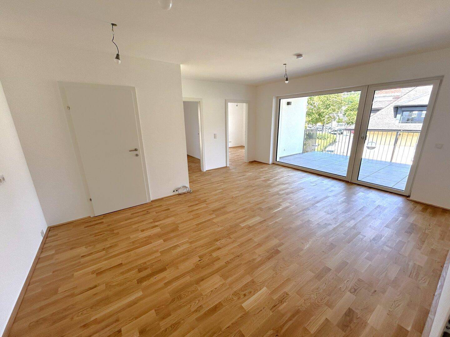 Helles, leeres Wohnzimmer mit warmem Parkettboden und direktem Zugang zu einem Balkon.