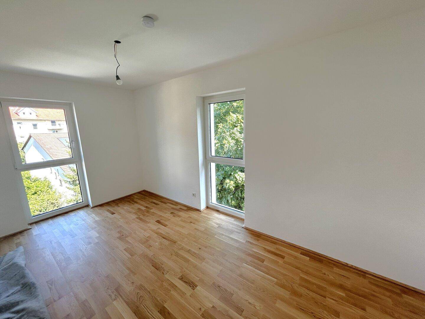 Helles Schlafzimmer mit Parkettboden und zwei Fenstern mit Blick ins Grüne.