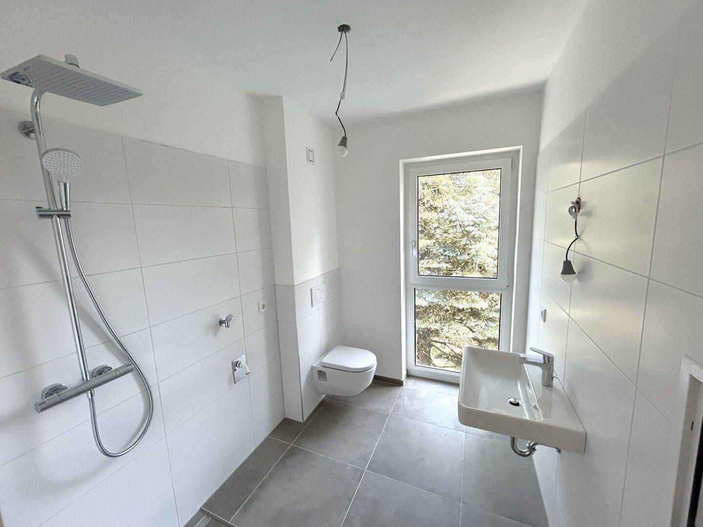 Modernes Badezimmer mit Dusche, WC und Fenster, hell gefliest in Weiß und Grau.