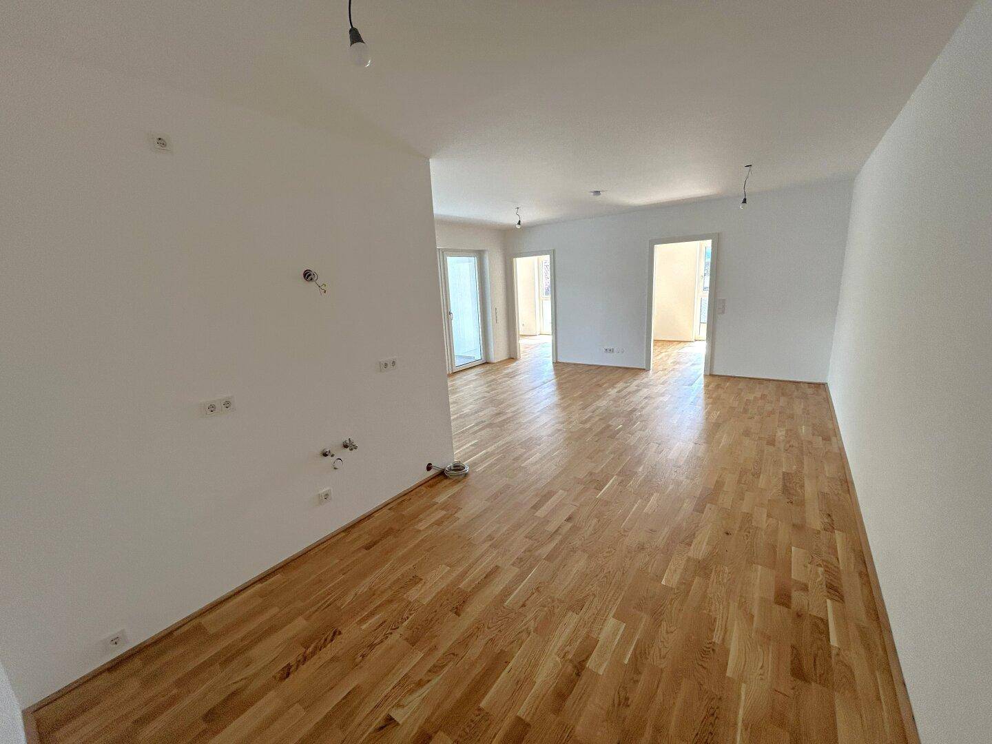 Geräumiges Wohnzimmer mit hellem Parkettboden und vorbereiteten Anschlüssen für Beleuchtung und Steckdosen.