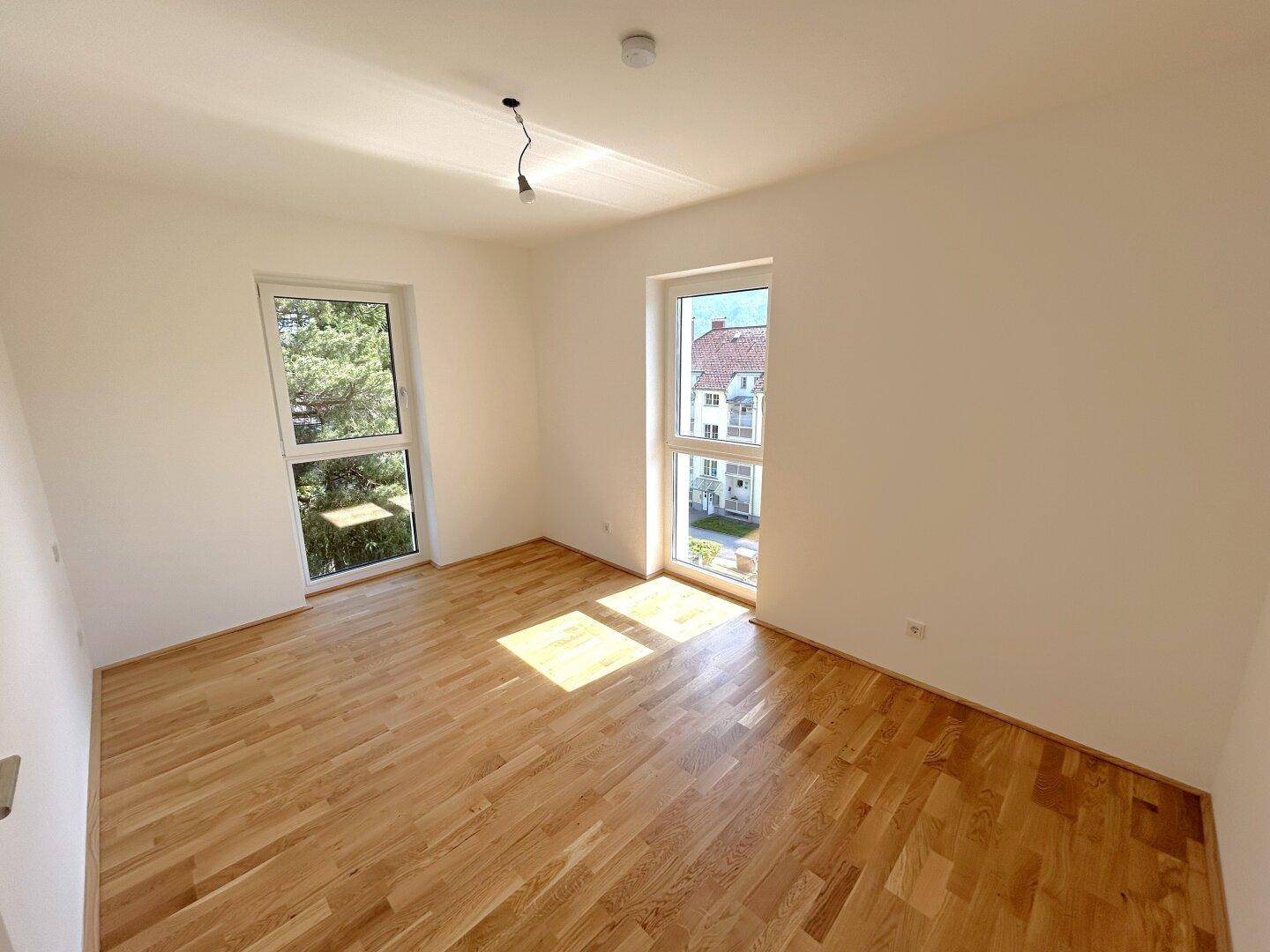Lichtdurchflutetes Schlafzimmer mit zwei Fenstern, hellem Parkettboden und weißen, frisch gestrichenen Wänden.