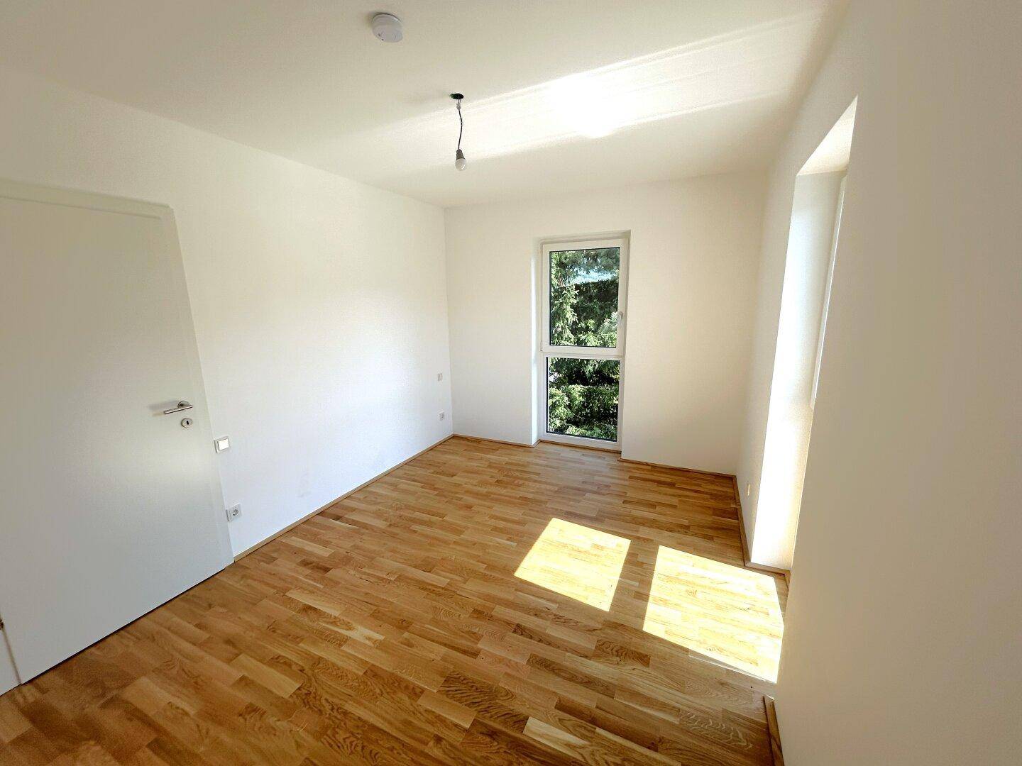 Geräumiges Schlafzimmer mit hellem Parkettboden, weißen Wänden und einem Fenster mit Blick ins Grüne.