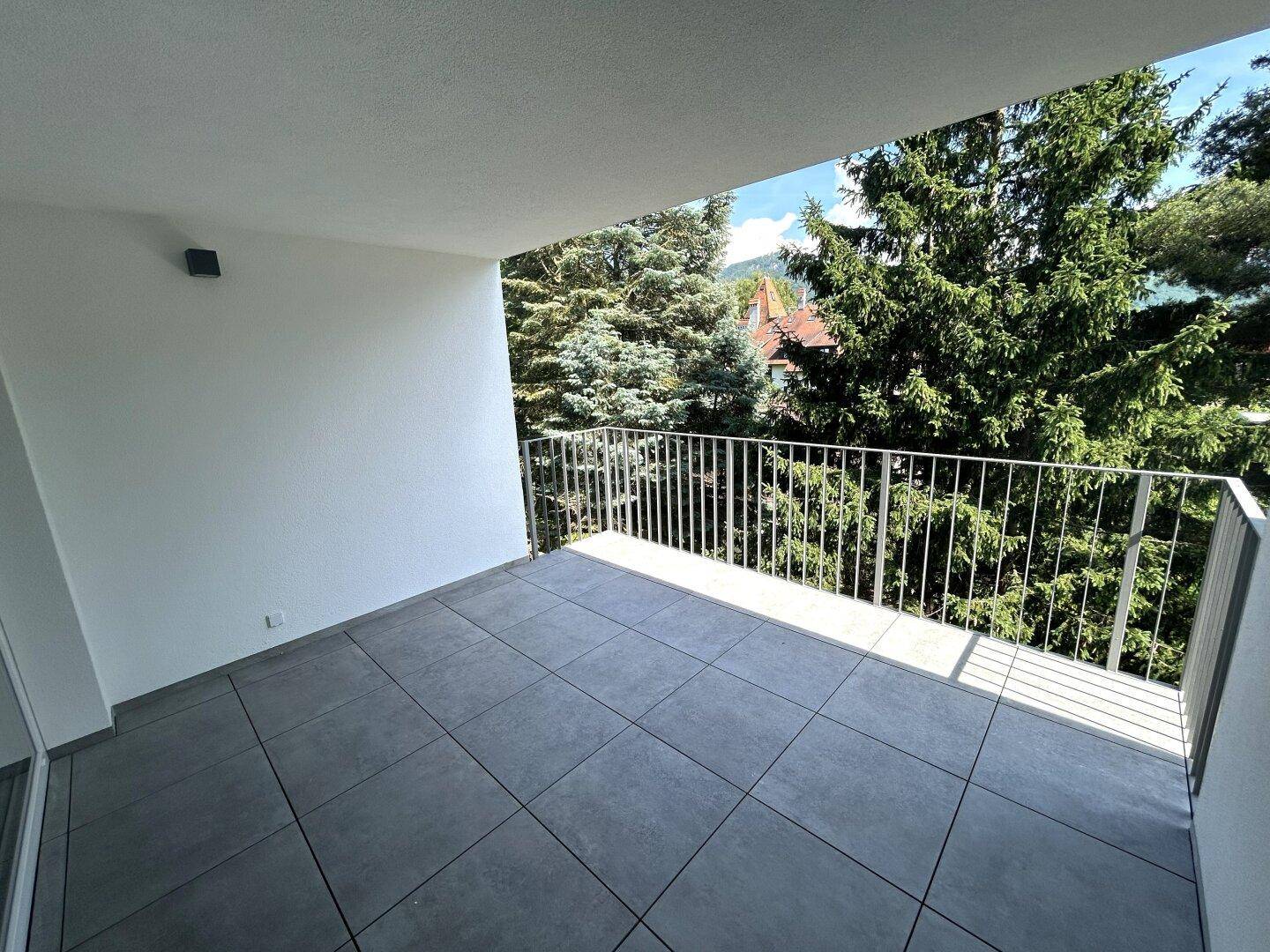 Sonniger Balkon mit Fliesenboden und Metallgeländer, umgeben von hohen Bäumen und viel Grün.
